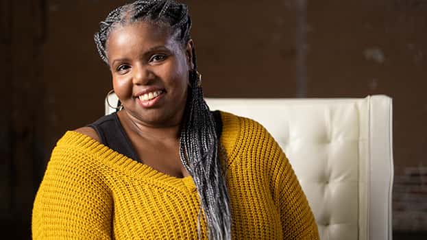 A smiling woman with braided hair, wearing a yellow sweater, sits on a white chair against a neutral background.