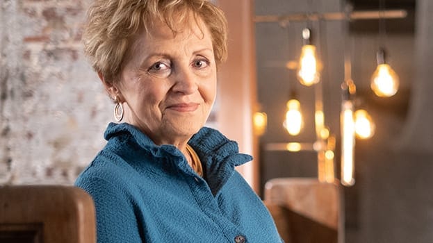 Older woman with short blonde hair, wearing a teal jacket, smiling gently while seated indoors with warm hanging lights in the background.