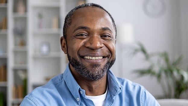 A middle-aged man with short hair and a salt-and-pepper beard, smiling warmly while wearing a light blue button-up shirt. He is sitting indoors with a blurred background of shelves and plants.